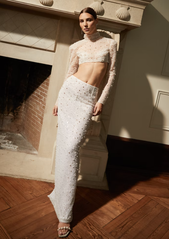 In questa foto una modella posa indossando un abito da sposa crop top, con mini top e gonna slim fit, tempestato di punti luce e micro perle della collezione autunno 2023 di Rita Vinieris