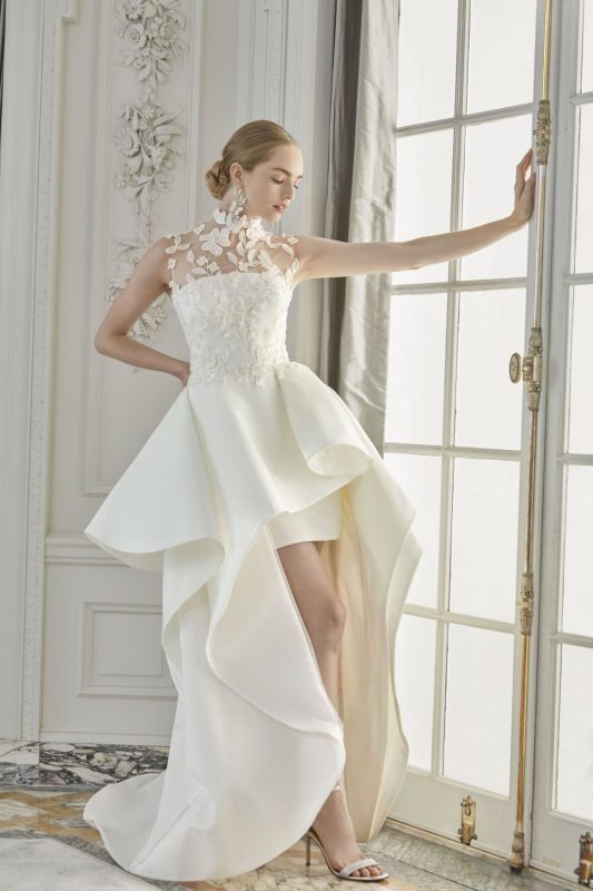 In questa foto una modella posa indossando un abito da sposa corto davanti e lungo dietro della collezione Sareh Nouri 2023