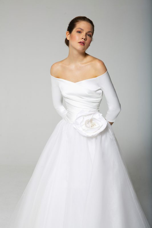 In questa foto una modella indossa un abito da sposa con scollo omerale e fiore maxi in vita della collezione Tosca Spose 2023