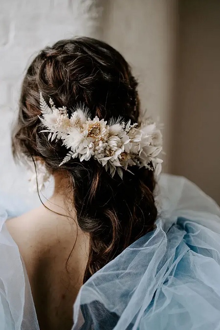 In questa foto, una sposa fotografata da dietro con un'acconciatura semi raccolta arricchita da una decorazione con fiori, foglie e piume