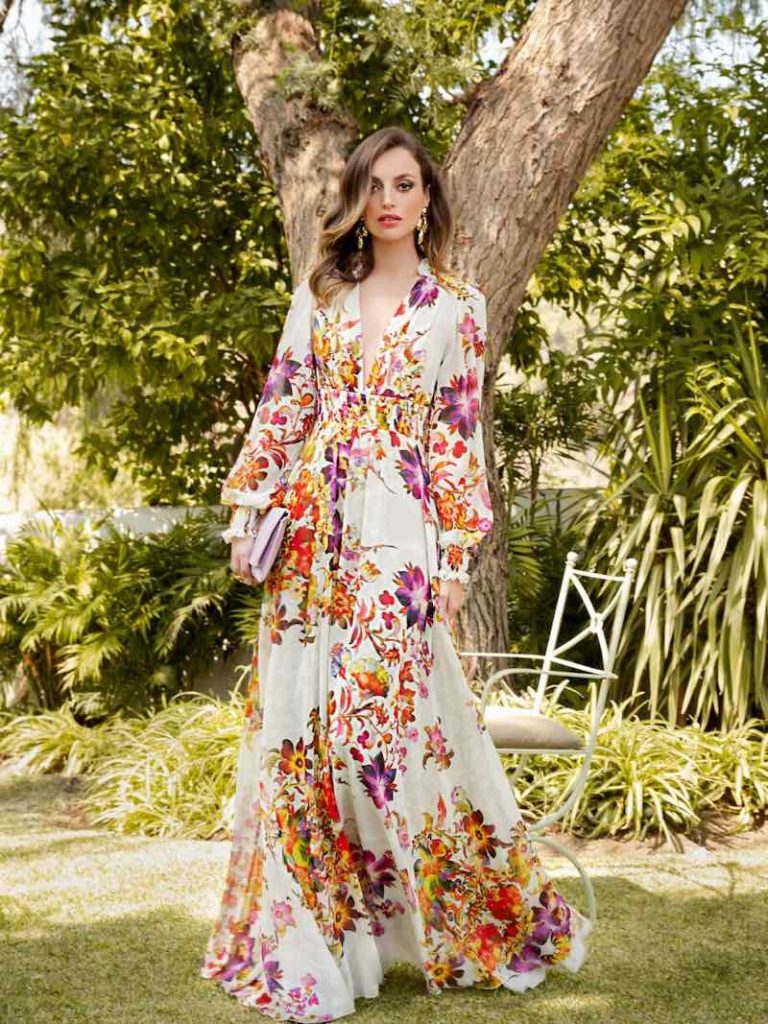 In questa foto una modella indossa uno degli abiti da cerimonia donna di Carla Ruiz, lunghi, con stampa floreale su fondo di colore bianco con  fiori di colore arancione, giallo, viola e rosso, maniche a sbuffo e scollo a V