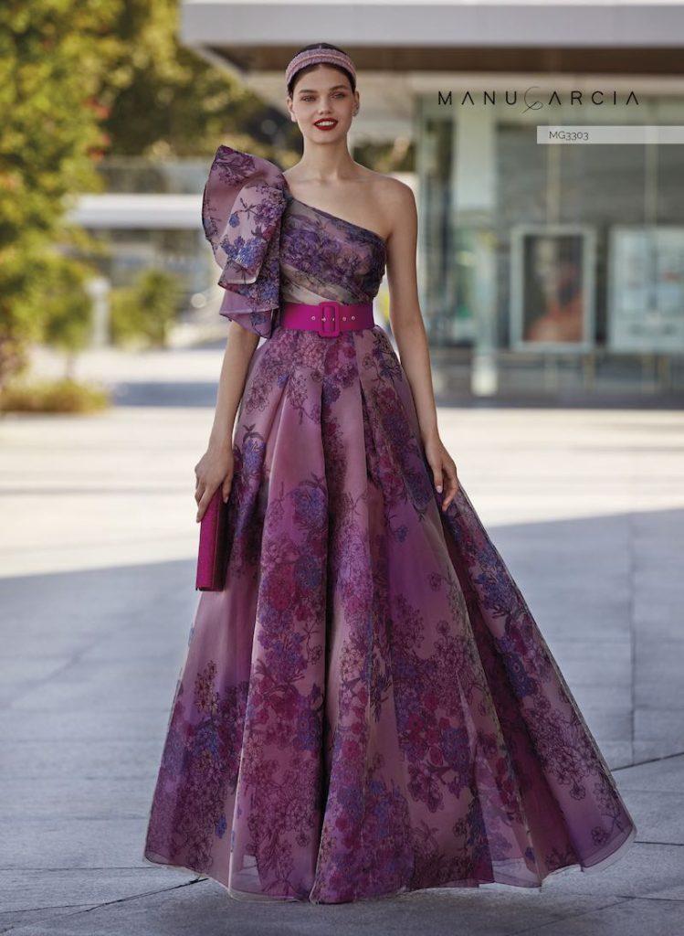 In questa foto una modella indossa un abito da cerimonia di Manu Garçia in tulle di colore rosa stampato con fiori viola con maxi fiocco monospalla e cintura di colore fucsia in vita