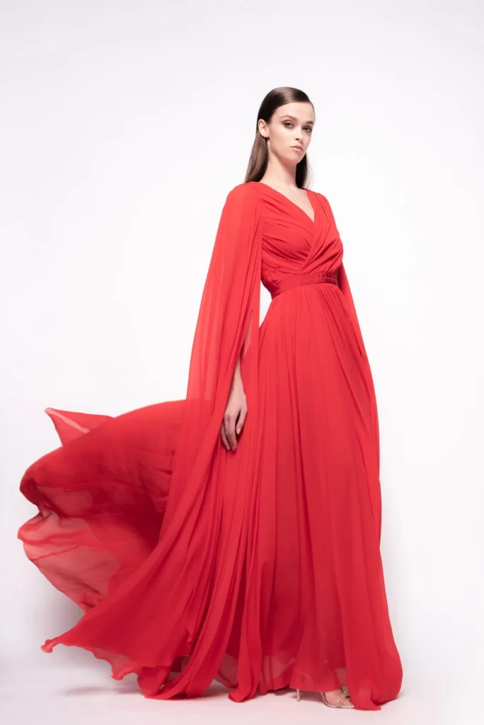 In questa foto una modella indossa uno degli abiti da cerimonia donna di Pinko lunghi, in chiffon di colore rosso aragosta, con drappeggio sul busto e maniche che si aprono come ali