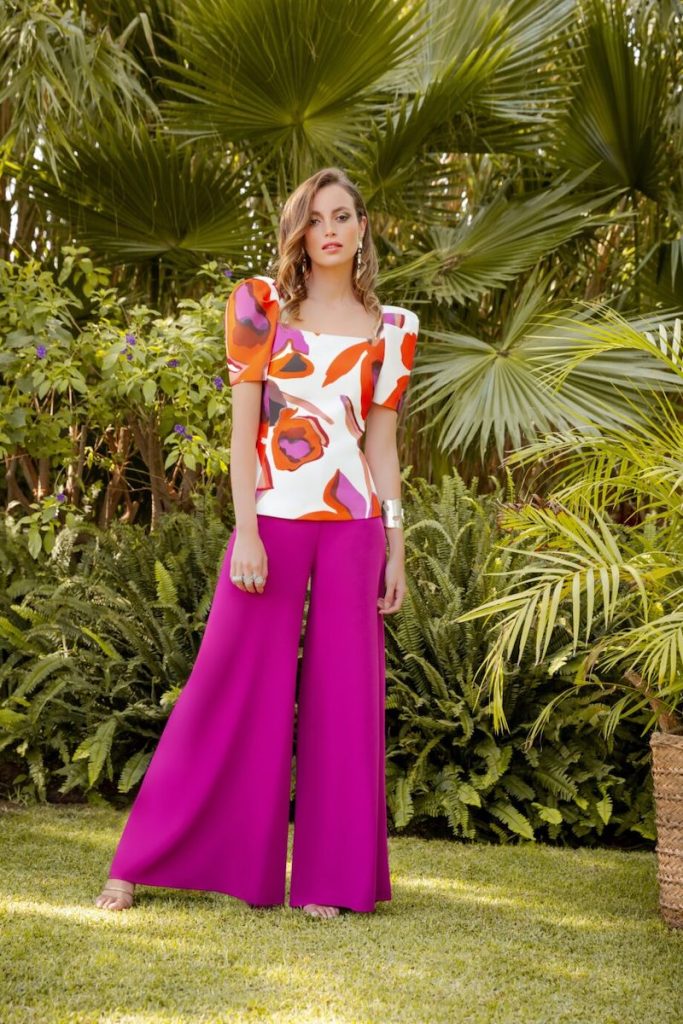 In questa foto una modella indossa uno degli abiti da cerimonia donna con pantaloni ampi di Carla Ruiz di colore fucsia con blusa sagomata con maniche corte a sbuffo e stampe floreali di colore arancione e rosa su sfondo di colore bianco