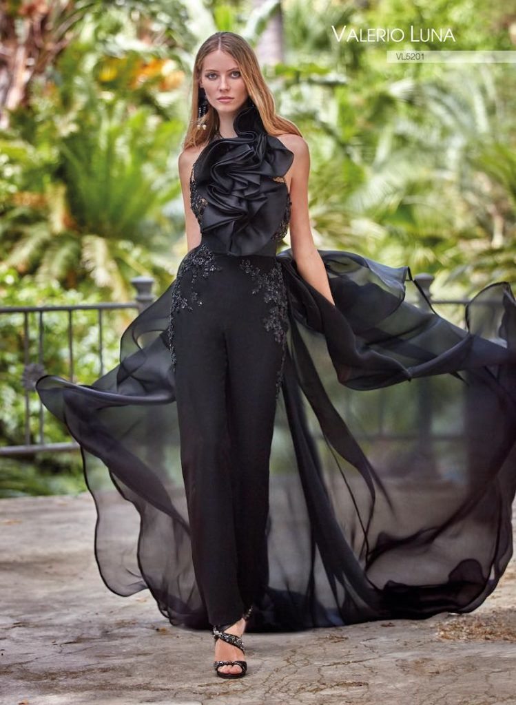 In questa foto una modella indossa una tuta elegante di colore nero con pantaloni leggermente ampi con ricami di perline e paillettes sui fianchi di Valerio Luna. Il corpetto si sviluppa con un grande fiore 3D. Dai pantaloni si allunga un ampio sovragonna di chiffon