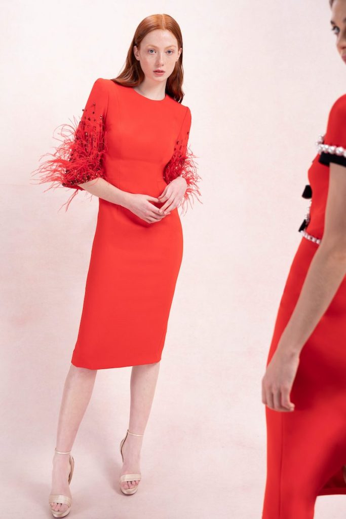 In questa foto una modella indossa uno degli abiti da cerimonia donna corti di Jenny Packham di colore rosso aragosta, con maniche a tre quarti e con piume sull'orlo delle maniche