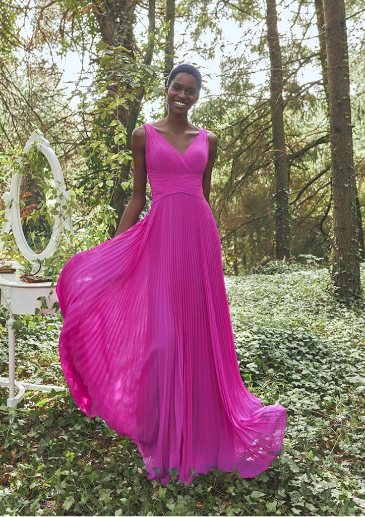 In questa foto una modella indossa uno degli abiti da cerimonia donna lunghi di colore fucsia di Pronovias. L'abito presenta un corpetto drappeggiato ad incrocio e una gonna scivolata plissé