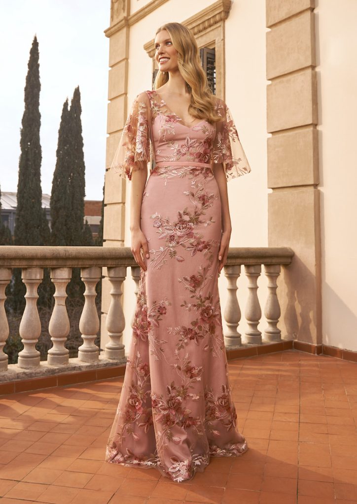 In questa foto una modella indossa uno degli abiti da cerimonia donna primaverili di Marchesa. Il modello è a semi sirena di colore rosa con stampe floreali e maniche corte in tulle ricamato