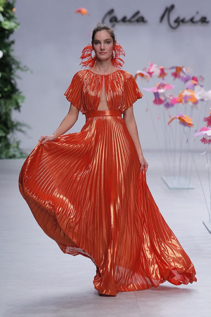 In questa foto una modella indossa uno degli abiti da cerimonia donna di Carla Ruiz in lurex di colore arancione. Il modello è lungo con tessuto plissé, con maniche corte e corpetto cut out 