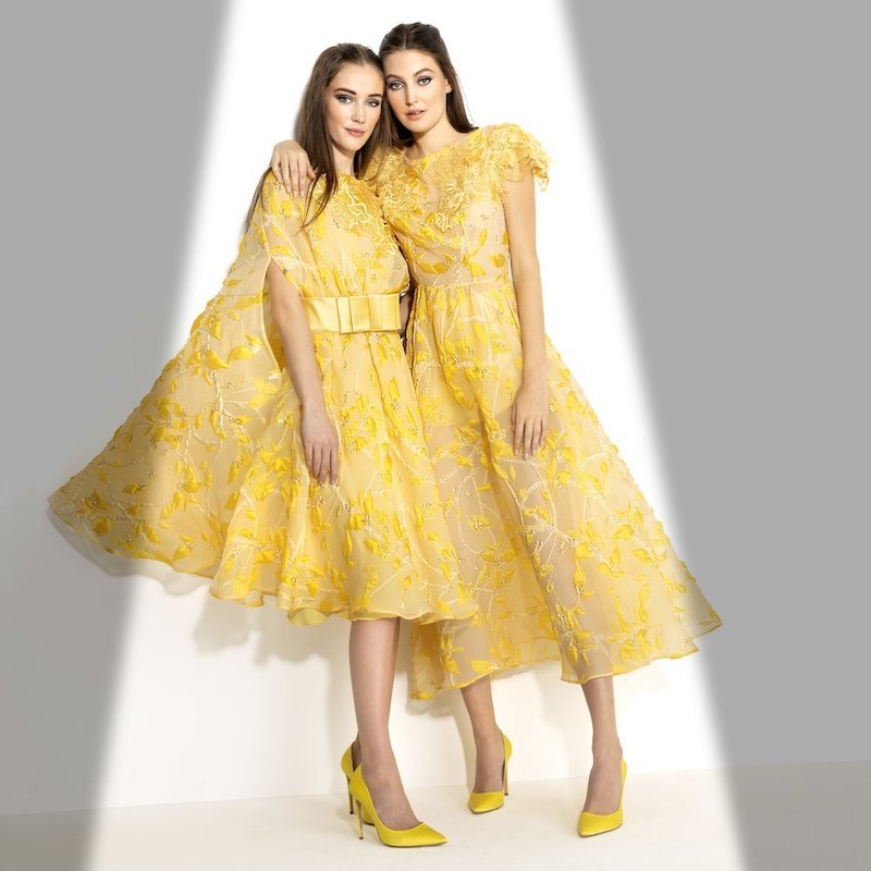 In questa foto due modelli indossano due abiti da cerimonia midi per testimone di Antonio Riva di colore giallo in chiffon con ricami jacquard all over. La modella a sinistra indossa anche un cinturone