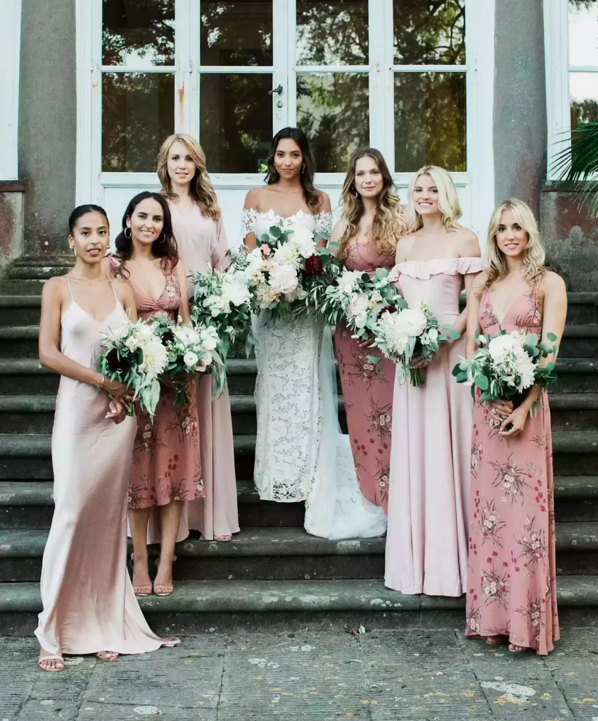 In questa foto una sposa posa sulle scale con le sue damigelle che indossano abiti da cerimonia donna di colore rosa e con stampe floreali. La sposa indossa un abito a sirena in pizzo. Tutte tengono tra le mani un bouquet di fiori bianchi e bordeaux