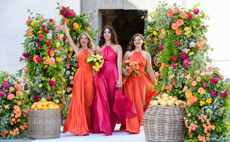 In questa foto tre ragazze indossano abiti da cerimonia donna per damigelle nello stesso modello con scollo all'americana ma in colori diversi: arancione e rosso fragola. Sono circondate da fiori degli stessi colori, da limoni e da arance riposti in due ceste di vimini