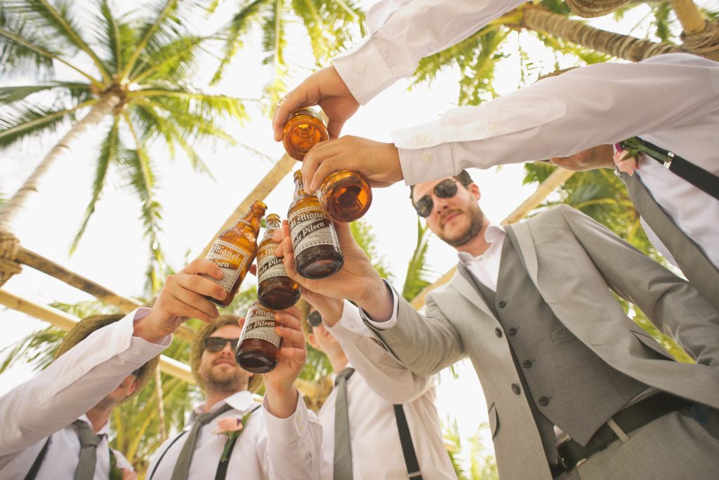 In questa foto scattata dal basso lo sposo brinda con i suoi amici con bottiglie di birra su una spiaggia tra le palme. Lo sposo indossa un abito grigio senza cravatta, gli amici una camicia con bretelle nere, cravatta grigia e cappelli di paglia