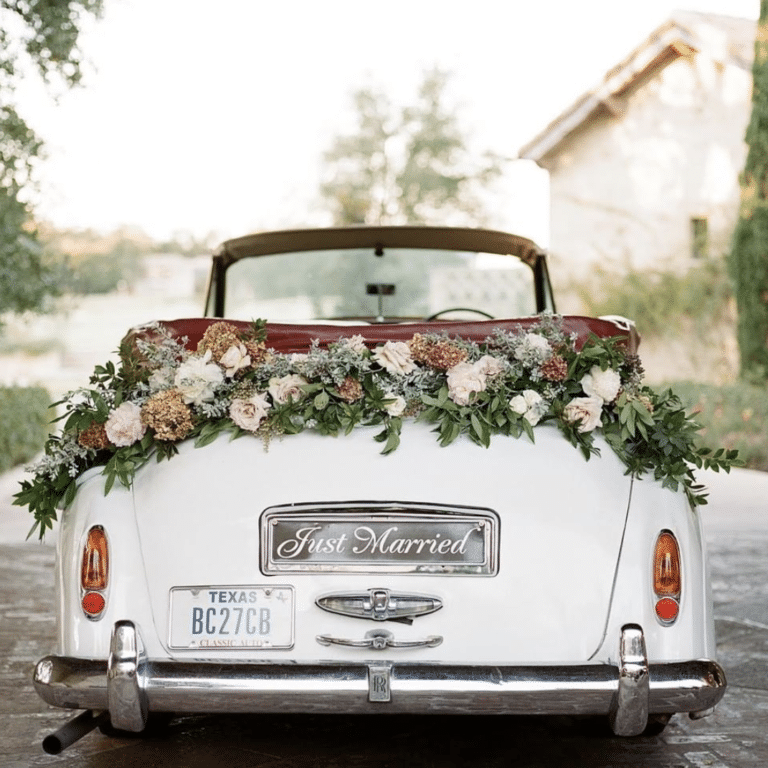 In questa foto un'auto per matrimonio  di colore bianco ritratta da dietro e decorata con un trancio di fiori e una targa con scritto "Just Married"