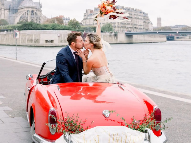 In questa foto due sposi lungo una riva del fiume si baciano a bordo della loro auto per matrimonio di colore rosso decorata con uno striscione con scritto "Just married". La sposa tiene in alto il suo bouquet nei colori del rosso e dell'arancione