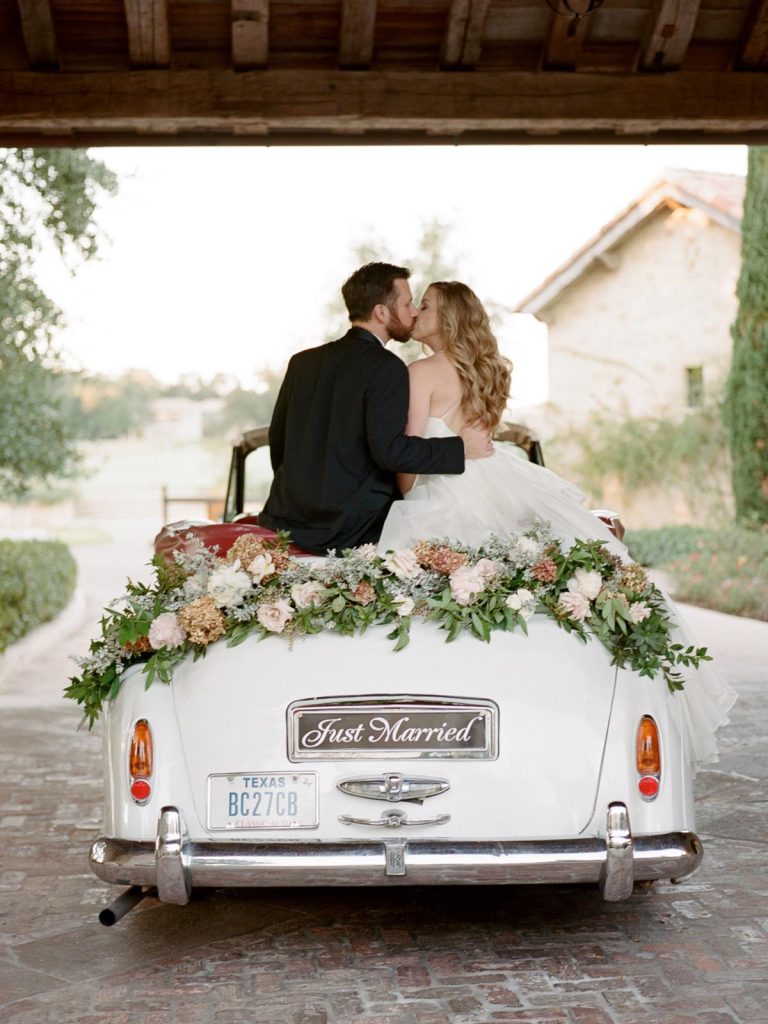 In questa foto due sposi a bordo della loro auto per matrimonio vintage ritratti di spalle mentre di baciano. L'auto è decorata con fiori e una targa personalizzata con scritto "Just Married" 