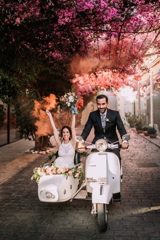 In questa foto due sposi su una Vespa vintage con sidecar di colore bianco decorata con fiori. La sposa tiene in mano il suo bouquet e un fumogeno di colore arancione