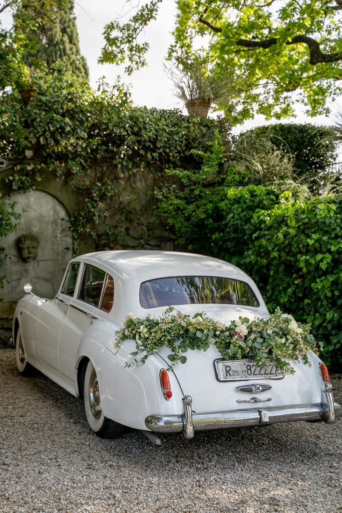 In questa foto un'auto per matrimonio  di colore bianco ritratta da dietro e decorata con un trancio di eucalipto