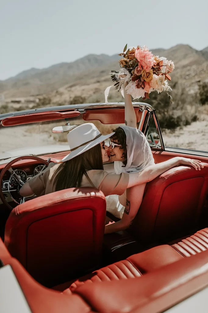 In questa foto due spose si baciano a bordo della loro auto per matrimonio decappottabile. Una sposa indossa un cappello texano bianco; l'altra sposa indossa un foulard 