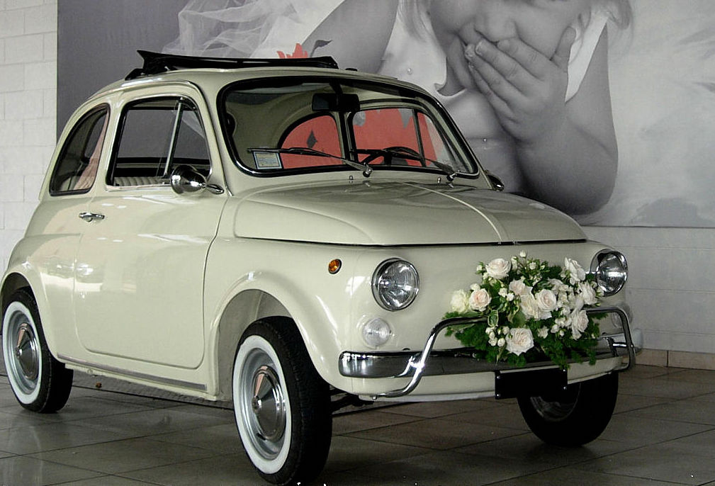 In questa foto una Fiat Cinquecento di colore bianco per matrimonio decorata con una composizione di rose di colore bianco sul paraurti anteriore