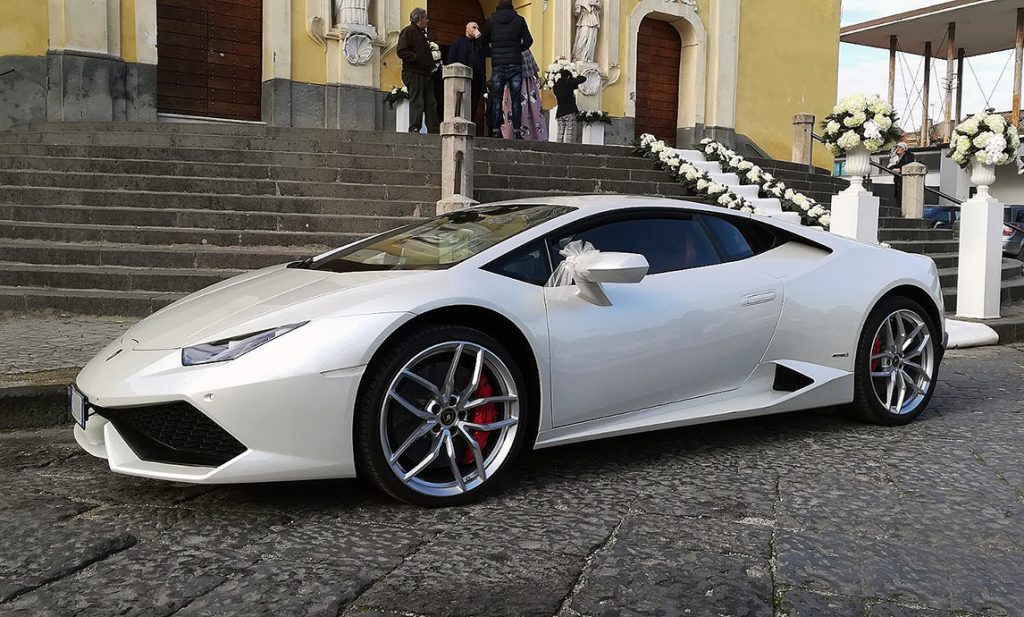 In questa foto una Lamborghini di colore bianco per matrimonio ritratta davanti alle scalinate di una chiesa e decorata con fiocchi di tulle sugli specchietti