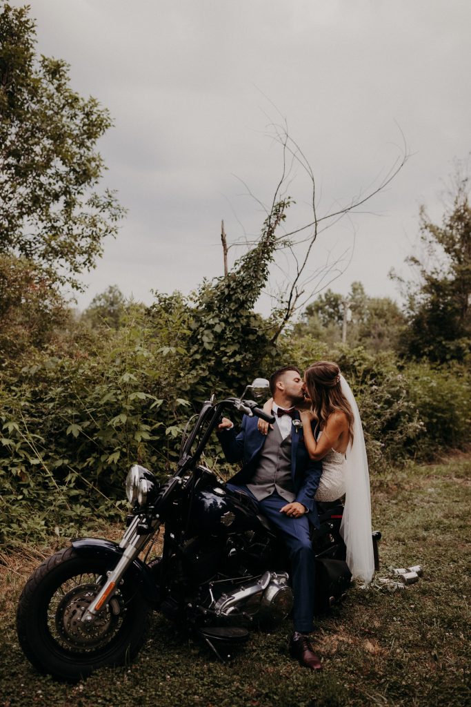 In questa foto due sposi si baciano a bordo di una motocicletta di colore nero parcheggiata su un prato