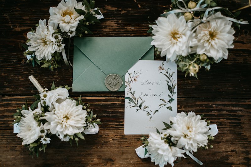 In questa foto una partecipazione di matrimonio con busta di colore verde bosco chiusa da ceralacca e card con il nome degli sposi circondate da dalie di colore bianco poggiate su un tavolo di legno