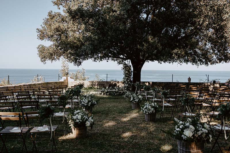 In questa foto la location di un matrimonio simbolico allestito sotto ad un grande albero decorato con un semi arco di rose e foglie. Lungo il corridoio centrale creato da due file di sedie campagnole sono presenti ceste di vimini con gli stessi fiori