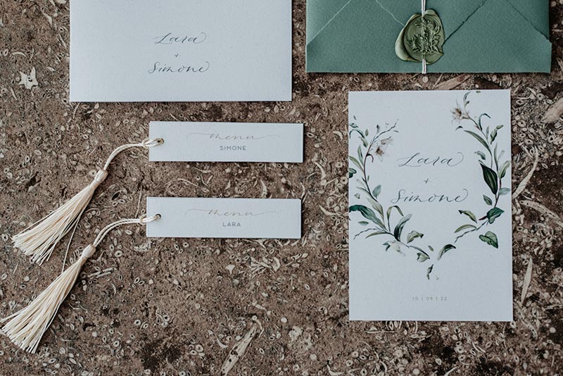 In questa foto un coordinato grafico  di matrimonio con busta della partecipazione di colore verde bosco chiusa da ceralacca, card con il nome degli sposi, libretto messa e menù-segnaposto con una nappina di colore bianco