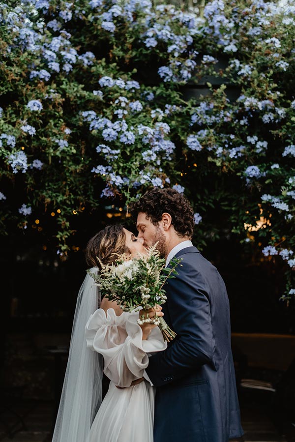 In questa foto due sposi si baciano sotto ad una pianta di glicine. La sposa tiene davanti al collo il suo bouquet di fiori di campo ed erbe aromatiche