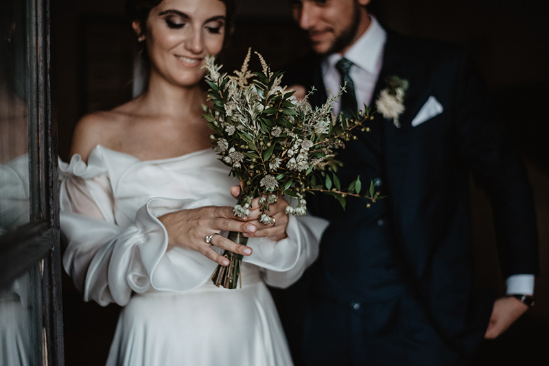 In questa foto una sposa in primo piano stringe e osserva sorridendo il suo bouquet di fiori di campo ed erbe aromatiche. Dietro di lei, lo sposo che la osserva sorridendo