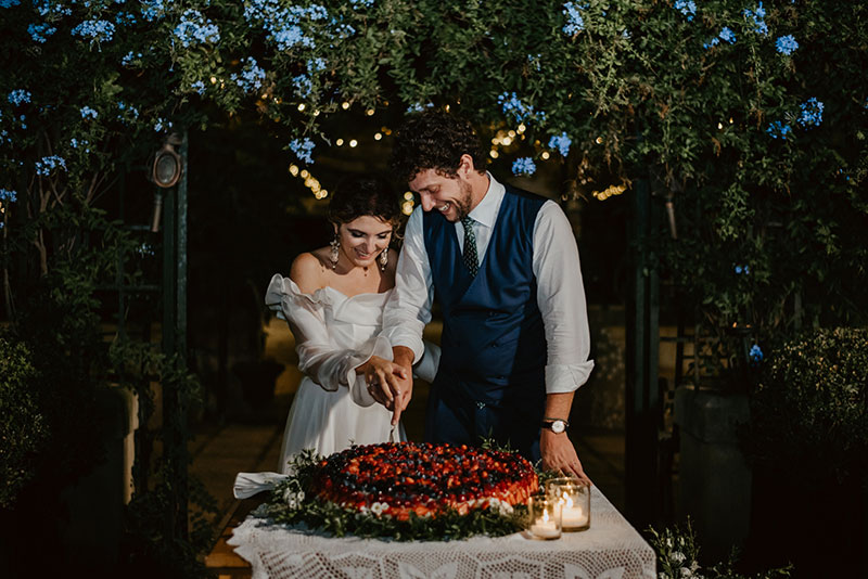 In questa foto due sposi, sorridendo, tagliano la loro torta nuziale ad un piano con fragoline e frutti di bosco
