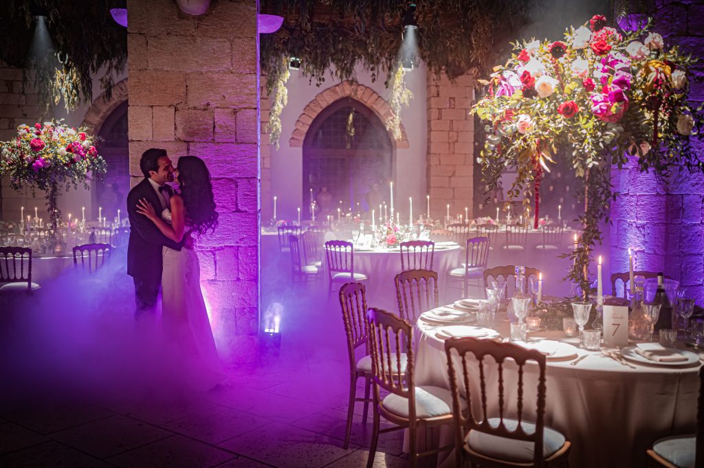 Gli sposi Carmen e Giuseppe ballano e si baciano tra le luci viola e il fumo nella sala del Castello Xirumi a Catania: organizzazione a cura della Wedding Planner Melania Millesi