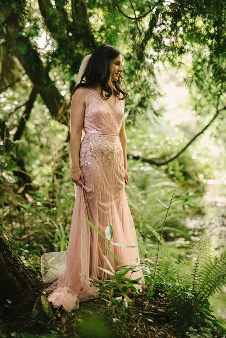 In questa foto, una sposa fotografata in un bosco, indossa un abito morbido in tulle rosa ricamato 