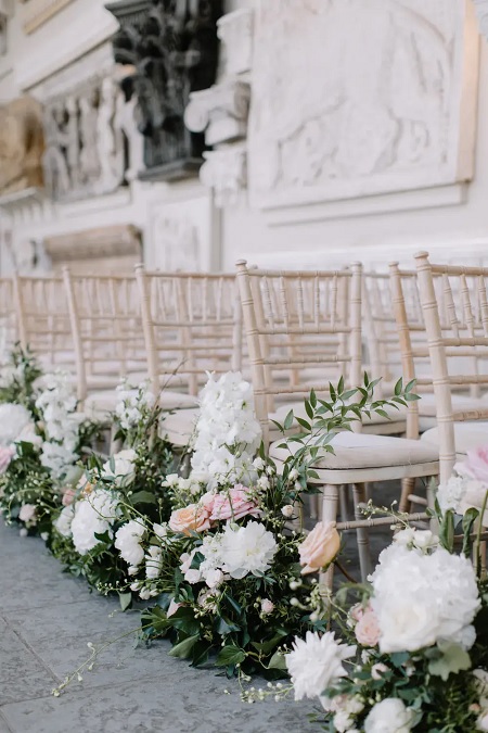 In questa foto, composizioni floreali con fiori bianchi, rosa e arancio chiari sistemati ai piedi di sedie color panna 