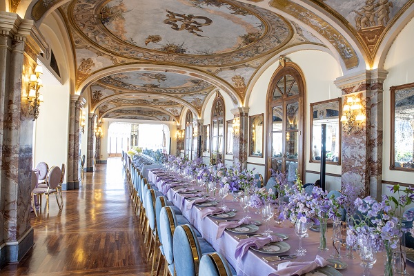 In questa foto, l'allestimento per il ricevimento di un matrimonio stile Bridgerton in una sala nobiliare nei toni del lilla e dell'azzurro polvere. La sala presenta affreschi sul soffitto con dettagli in colore oro. Le sedie utilizzate sono in velluto con dettagli in colore oro. Sul tavolo sono presenti vasi di vetro con fiori di colore lilla e tovaglioli abbinati poggiati su piatti in ceramica