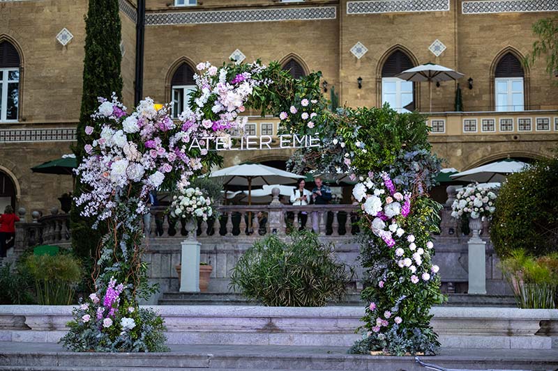 In questa foto un arco di fiori di colore rosa e bianco con scritta luminosa Atelier Emé davanti alla facciata di Villa Igiea