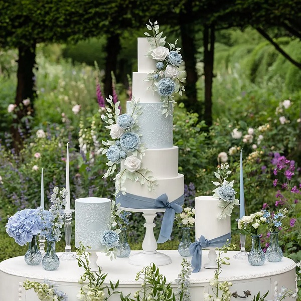 In questa foto, il tavolo con la torta nuziale a più piani bianchi e azzurri e due torte più piccole è allestito all'esterno e arricchito con fiori e candele 