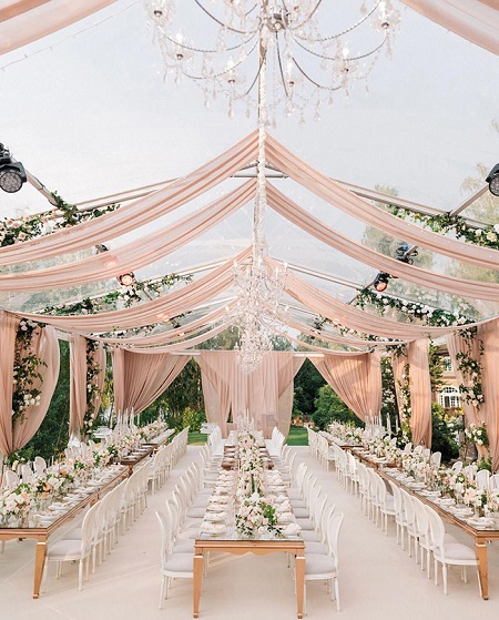 In questa foto, un ricevimento di nozze all'aperto con tre tavoli imperiali disposti in verticale decorati con fiori bianchi e rosa e disposti sotto tende rosa brillante