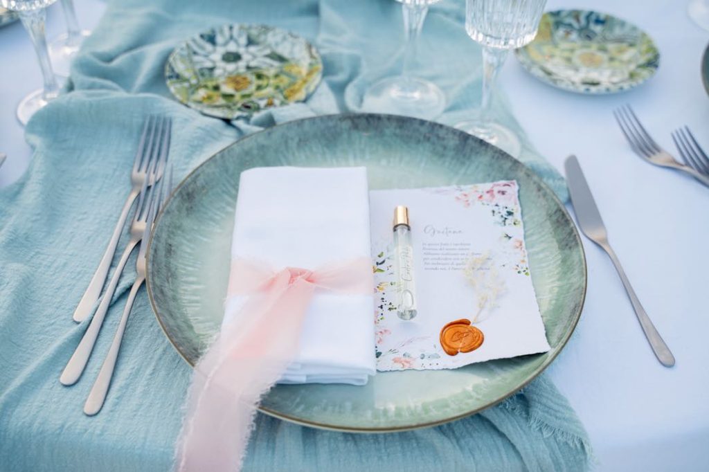 In questa foto un dettaglio della mise en place: il piatto segnaposto verde, posizionato su un runner di lino in verde salvia, ospita il tovagliolo legato con un nastrino di chiffon rosa e il segnaposto con il profumo personalizzato omaggio degli sposi ai loro ospiti