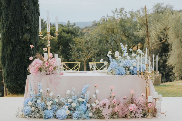 In questa foto, il tavolo degli sposi all'esterno è decorato con composizioni di fiori bianchi, azzurri e rosa poggiati a terra e sul tavolo, arricchiti con candelabri dorati