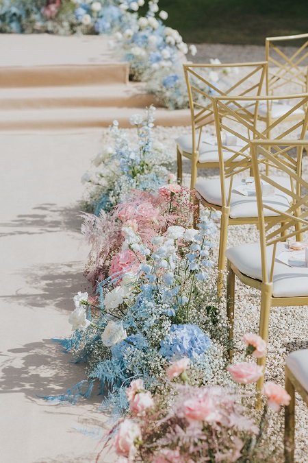 In questa foto, composizioni di fiori bianchi, azzurri e rosa sono sistemate a terra vicino alle sedie degli invitati in una cerimonia all'aperto