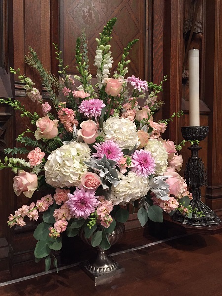 In questa foto, una composizione di rose e delphinium rosa cipria, gerbere rosa brillante e ortensie bianche sistemata all'interno di un vaso d'argento 