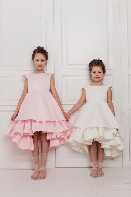 In questa foto, due bambine fotografate di fronte si tengono per mano. La maggiore indossa un abito rosa brillante, la minore bianco, i piedi nudi sono coperti con calzari di tulle a puntini 
