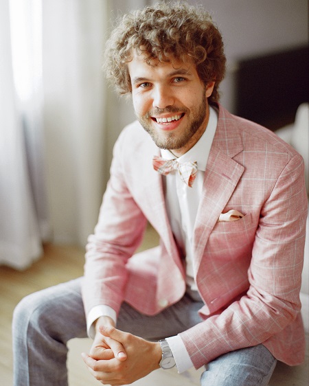 In questa foto, uno sposo seduto indossa un abito spezzato con pantaloni grigi, camicia bianca, giacca, papillon e fazzoletto da taschino in rosa brillante 