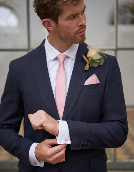 In questa foto, uno sposo fotografato a mezzo busto indossa un abito blu scuro, cravatta, fazzoletto da taschino e gemelli in rosa brillante, colore della Biodiversità di Pantone 