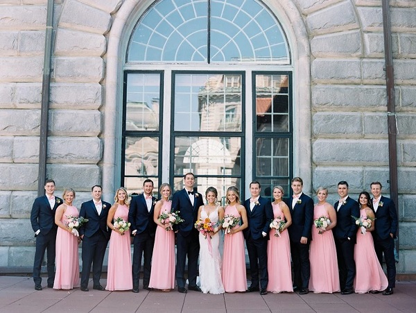 In questa foto, uno scatto di gruppo con al centro la sposa in abito bianco, lo sposo in abito e papillon blu scuro, ai lati tre coppie di damigelle adulte in abito rosa brillante e accompagnatori in abito scuro e papillon rosa 