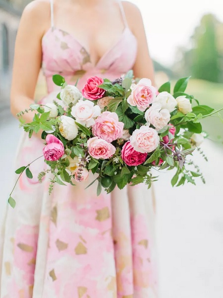 In questa foto, si intravede una sposa che indossa un abito floreale sui toni del rosa e del verde, con in mano un bouquet di rose dai toni del rosa chiaro e scuro