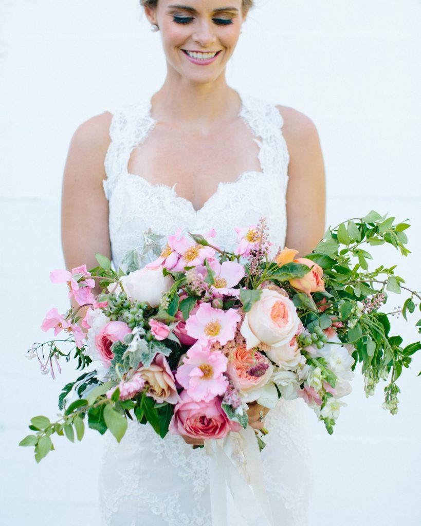 In questa foto, una sposa in abito bianco tiene in mano un bouquet scomposto di peonie, rose da giardino, dalie, gelsomino e ranuncolo