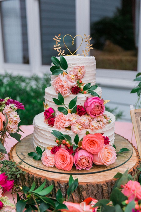 In questa foto, una torta floreale a più piani poggiata su un piatto di vetro e la fetta di un tronco d'albero, con glassa bianca e mix di fiori rosa brillante 